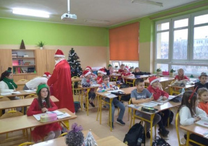 Na zdjęciu widać uczniów w sali lekcyjnej oraz Mikołaja wręczającego prezenty.