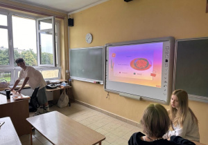 Na zdjęciu widać uczniów klas siódmych podczas zajęć interaktywnych z języka niemieckiego.Uczeń stojąc przy biurku wyświetla prezentacje na tablicy interaktywnej.
