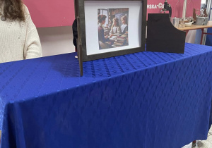 Na zdjęciu widać stół, na którym prezentowane jest zdjęcie wygenerowane przez stuczną inteligencję. Za stołem siedzi uczennica - uczestniczka uroczystej akademii.