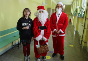 Na zdjęciu widać stojących na korytarzu szkolnym dwóch Mikołajów w towarzystwie Śnieżynki. Jeden z nich trzyma kosz pełen słodkości.