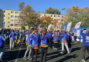 Na zdjęciu widzć uczestników gier sportowych stojących na boisku szkolnym przed przystąpieniem do wykonywania zadań, podczas wspólnej rozgrzewki.