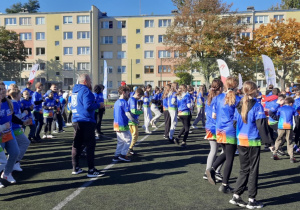 Na zdjęciu widzć uczestników gier sportowych stojących na boisku szkolnym przed przystąpieniem do wykonywania zadań podczas wspólnej rozgrzewki.