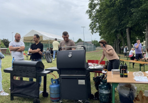 Trzech mężczyzn obsługujących dwa grille gazowe na trawie, przygotowujących jedzenie dla uczestników festynu rodzinnego.