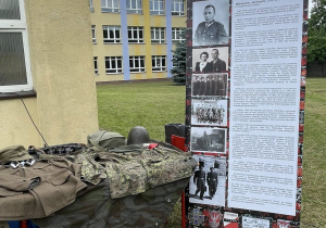 Stoisko edukacyjne z roll-upem poświęconym Kapitanowi Władysławowi Jachowiczowi 'Konar' i eksponatami wojskowymi, na tle budynku szkolnego.