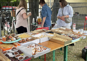 Stoisko z dużą ilością domowych ciast, babeczek i ciasteczek w jednorazowych pojemnikach, obsługiwane przez dorosłe kobiety na festynie szkolnym.
