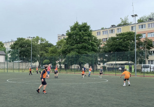 Uczniowie w pomarańczowych kamizelkach grający w piłkę nożną na sztucznej murawie boiska szkolnego podczas festynu.