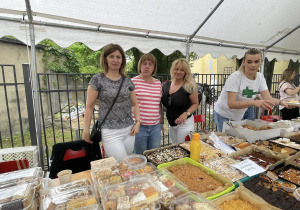 Cztery kobiety za stoiskiem z licznymi domowymi ciastami i słodkościami, przygotowanymi na sprzedaż podczas festynu rodzinnego.