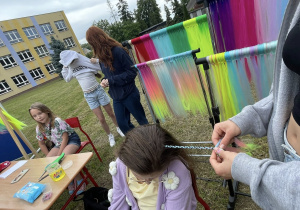 Dziewczynka siedząca na krześle, której ręce inna osoba zaplata w kolorowe warkoczyki z syntetycznych włosów, na tle stoiska z długimi, kolorowymi pasmami