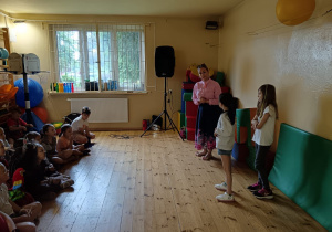 Nauczycielka stoi z dwoma uczennicami na środku sali z drewnianą podłogą, rozmawiając z dużą grupą dzieci siedzących na podłodze. W tle widać głośnik i okno.