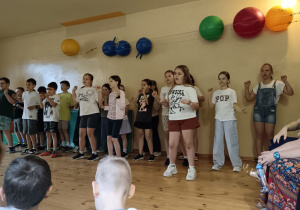 Grupa około dziesięciu dzieci stojących w rzędzie na scenie lub w sali, śpiewająca i wykonująca gesty rękami w rytm muzyki. W tle widać kolorowe ozdoby ścienne.