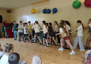 Grupa dzieci stojących w linii, trzymających się za ręce lub dotykających ramion w tańcu lub grze, na drewnianej podłodze sali. W tle kolorowe ozdoby ścienne.
