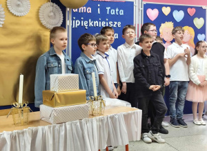 Grupa około dziesięciu chłopców w eleganckich strojach, stojących na tle niebieskiej tablicy z napisem 'Mama i Tata' i ozdobami w kształcie serc, przygotowujących się do występu.