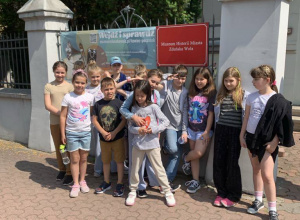 Uczniowie klasy 2b pozują przed wejściem do Muzeum Historii Miasta Zduńska Wola, obok bramy z szyldem 'Wędź i sprawdź' i 'Muzeum Historii Miasta Zduńska Wola'. Dzień jest słoneczny.