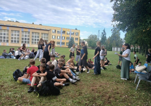 Na zdjęciu uczniowie klas 7 i 8 wraz z opiekunami, na boisku szkolnym, podczas czytania fraszek Jana Kochanowskiego.