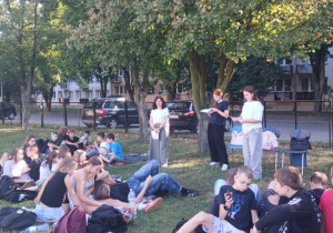 Na zdjęciu uczniowie klas 7 i 8 wraz z opiekunami, na boisku szkolnym, podczas czytania fraszek Jana Kochanowskiego.