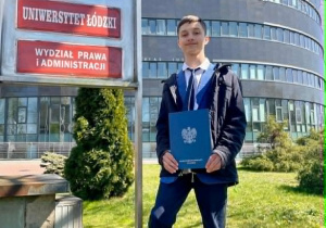 Na zdjęciu Arkadiusz, z pamiątkowym dyplomem laureata, stoi przed budynkiem Wydziału Prawa i Administracji Uniwersytetu Łódzkiego.