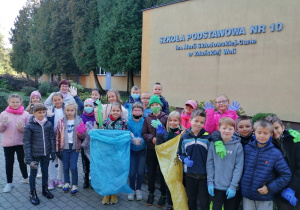 Uczniowie na tle budynku szkolnego, ubrani w rękawice ochronne szykują się do sprzątania placu wokół boiska szkolnego.