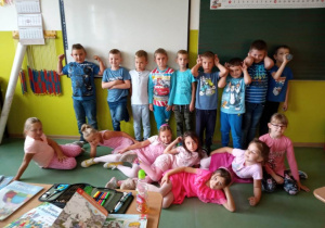 Zdjęcie uczniów klas 1 - 3 w izbie lekcyjnej. Część uczniów siedzi na podłodze, część natomiast stoi. Dziewczynki ubrane w kolory różowe, chłopcy w stroje niebieskie.