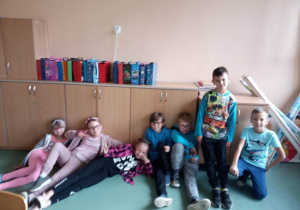 Zdjęcie uczniów klas 1 - 3 w izbie lekcyjnej. Część uczniów siedzi na podłodze, część natomiast stoi. Dziewczynki ubrane w kolory różowe, chłopcy w stroje niebieskie.