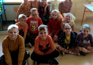 Na zdjęciu widać siedzące w klasie na podłodze uśmiechnięte dzieci.