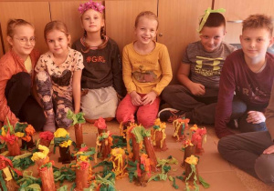 Uczniowie siedząc prezentują wykonane przez siebie prace. Są to wykonane z papieru drzewka w barwach jesieni.