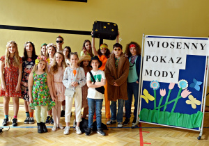 Na zdjęciu widać wszystkich uczestników konkursu pokazu mody. Uczniowie stoją na sali gimnastycznej obok parawanu z napisem : "WIOSENNY POKAZ MODY".