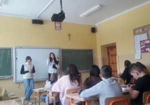 Na zdjęciu widać siedzących w pracowni uczniów oraz stojące na tle tablicy dwie uczennice prowadzące zajęcia dla swoich kolegów i koleżanek z klasy.