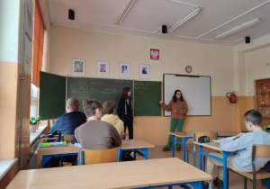 Na zdjęciu widać siedzących w klasie uczniów, dwie uczennice zajęły miejsce nauczyciela i prowadzą zajęcia.