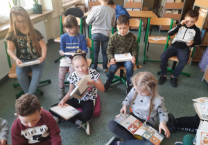 Dzieci z klas pierwszych siedzą na podłodze w szkolnej bibliotece i przeglądają książki..