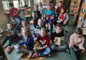 Dzieci z klas pierwszych siedzą na podłodze w szkolnej bibliotece i przeglądają książki.