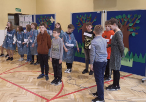 Na zdjęciu widać uczniów z klasy 5a podczas występu. Uczniowie stoją w sali gimnastycznej na tle dekoracji z napisem "Dzień Ziemi".