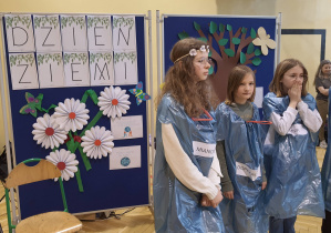 Na zdjęciu widać uczniów z klasy 5a podczas występu. Uczniowie stoją w sali gimnastycznej na tle dekoracji z napisem "Dzień Ziemi".