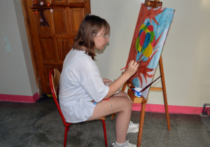 Uczennica siedzi przy sztaludze i maluje obraz.