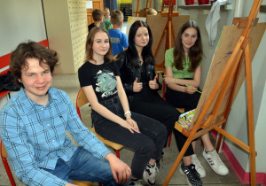 Uczniowie z klas starszych, podczas malowania obrazu, pozują przy sztaludze.