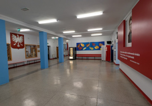 Zdjęcie przedstawia dużą, przestronną halę lub rozwidlenie korytarzy w szkole. Podłoga jest jasnoszara, a ściany i sufit białe. Centralnym elementem są trzy solidne, kwadratowe kolumny pomalowane na jaskrawy, jasnoniebieski kolor. Po lewej stronie widać wiszące, duże, biało-czerwone godło państwowe – polskiego Orła Białego – na tle czerwonego tła. Poniżej znajdują się gabloty z trofeami. Po prawej stronie dominuje duży, czerwony panel ścienny, na którym widoczny jest portret i inspirujący cytat. Na wprost, między kolumnami, znajduje się czarny automat vendingowy. W tle, na ścianie, widoczny jest barwny mural z motywem jesiennych liści w odcieniach żółci, czerwieni i brązu. Wzdłuż ścian ciągną się proste, niskie, czerwone ławki.