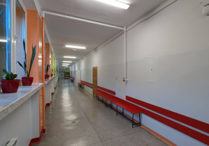 Zdjęcie ukazuje długi i prosty korytarz, charakteryzujący się doskonałym oświetleniem. Wzdłuż lewej ściany biegnie rząd dużych okien, wpuszczających dużo światła dziennego. Parapet okienny ma jasnopomarańczowy kolor. Na parapecie regularnie ustawiono rośliny doniczkowe (prawdopodobnie sansewierie) w jaskrawoczerwonych doniczkach, ożywiające przestrzeń. Po prawej stronie wzdłuż białej ściany ciągną się czerwone listwy ochronne i rząd długich, czerwonych ławek. Korytarz ma jasną podłogę i biały sufit, co potęguje wrażenie przestronności.