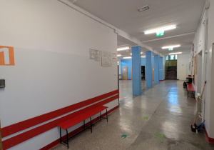 Kadr przedstawia wejście do strefy korytarzowej. Po lewej stronie widać drewniane drzwi wejściowe, obok których na białej ścianie znajduje się duże, pomarańczowe oznaczenie z cyfrą "1". Pod drzwiami zainstalowany jest panel dostępu (np. czytnik kodów). Wzdłuż ściany ciągną się niskie, czerwone ławki oraz czerwone listwy ochronne. Korytarz wiedzie prosto w głąb, gdzie widać cztery jasnoniebieskie kolumny. Na końcu korytarza, po prawej stronie, widać wejście na schody. Na podłodze widoczne są zielone naklejki kierunkowe w formie strzałek lub śladów stóp.